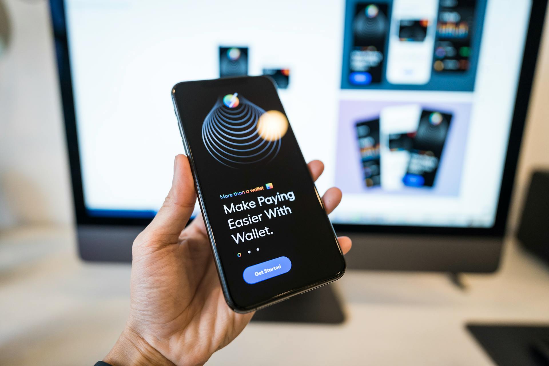 Hand holding smartphone displaying digital wallet app interface, blurred monitor in background.