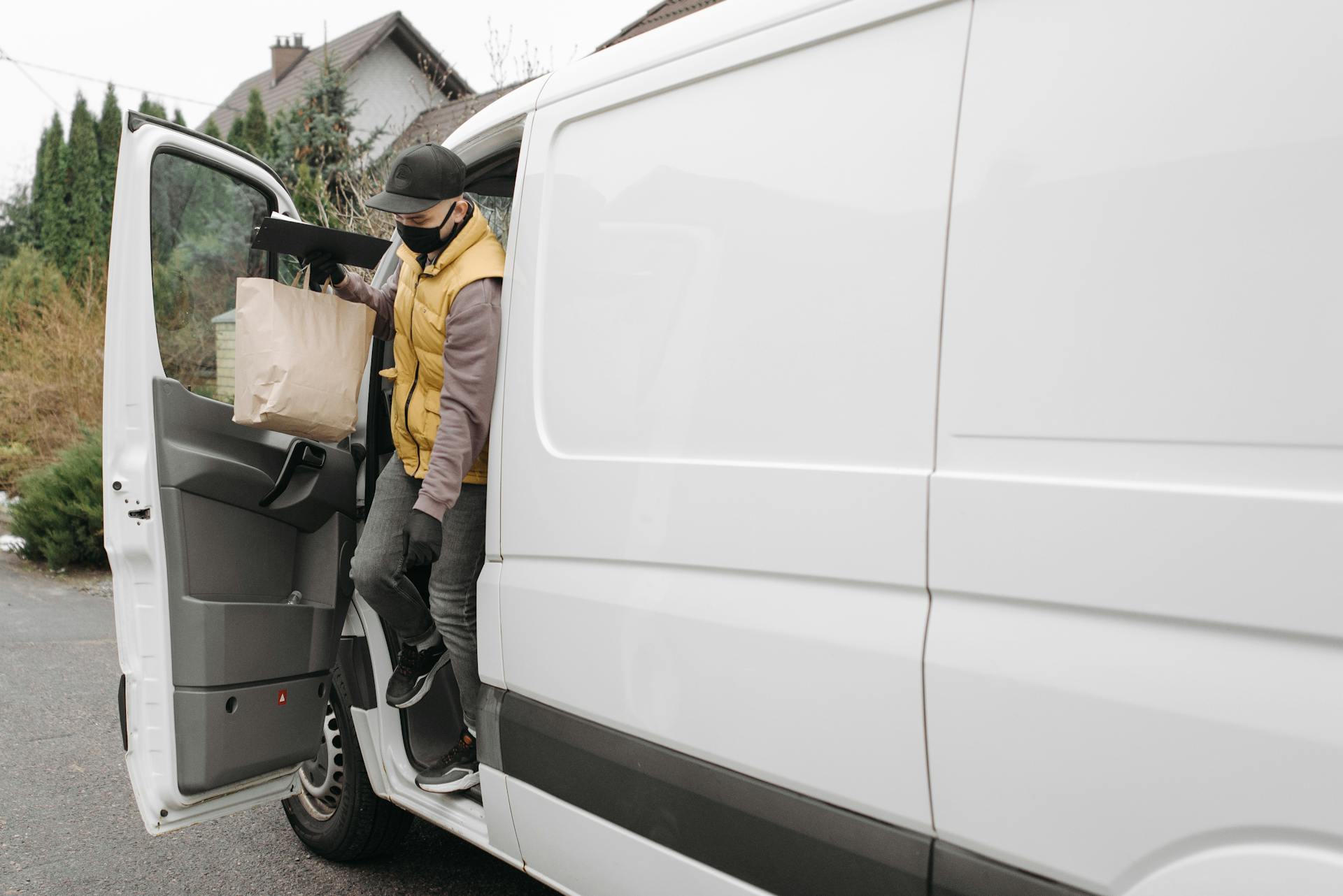 Masked delivery man exits van holding paper bag, showcasing modern delivery service.