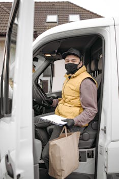 A delivery driver wearing a face mask sits inside a van holding a paper bag and clipboard, symbolizing contactless delivery