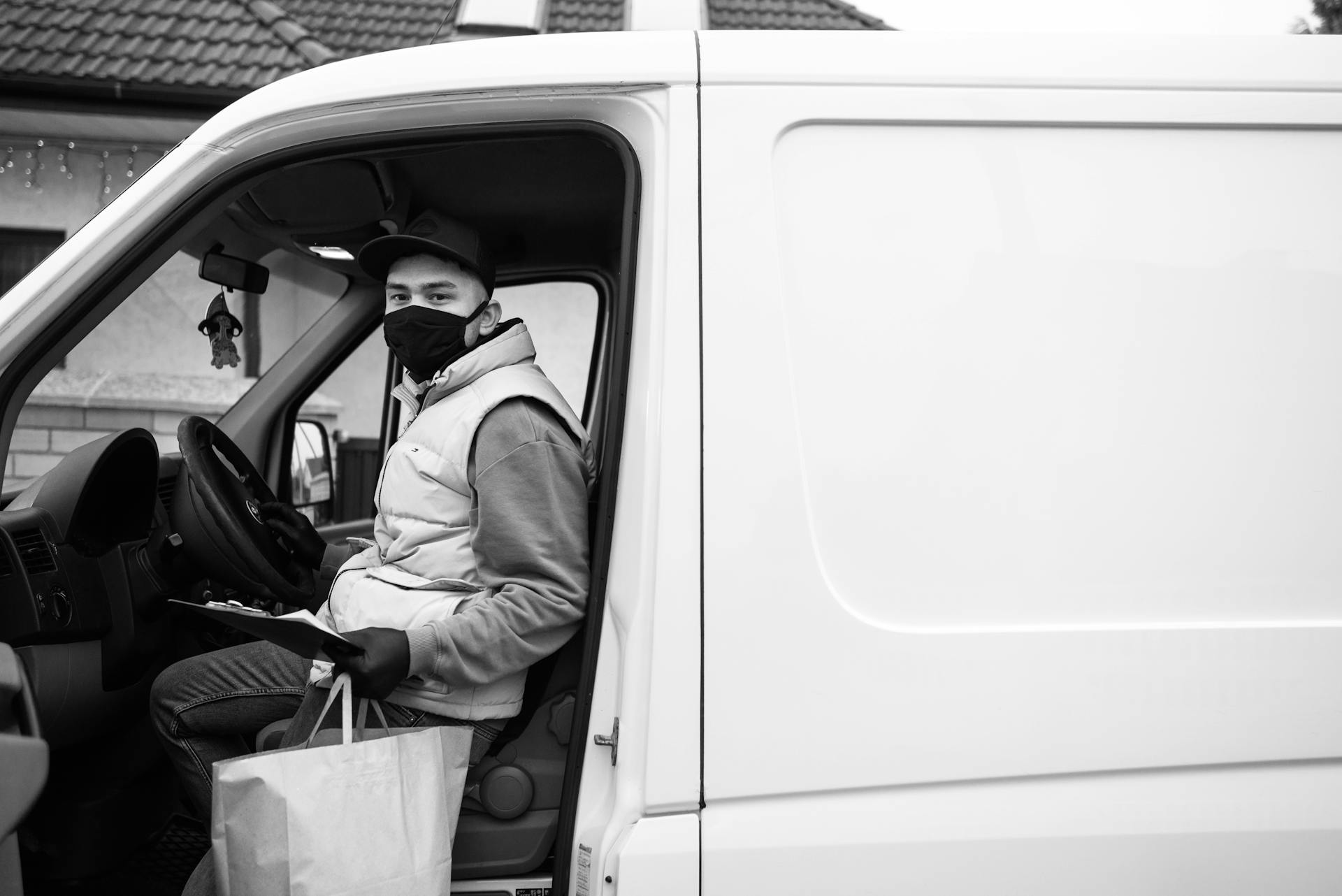 Black and white image of a masked delivery driver in a van, highlighting safety measures.