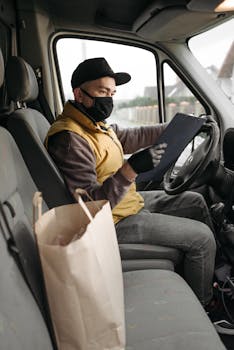 A delivery person wearing a mask holds a clipboard inside a vehicle, preparing orders for delivery.