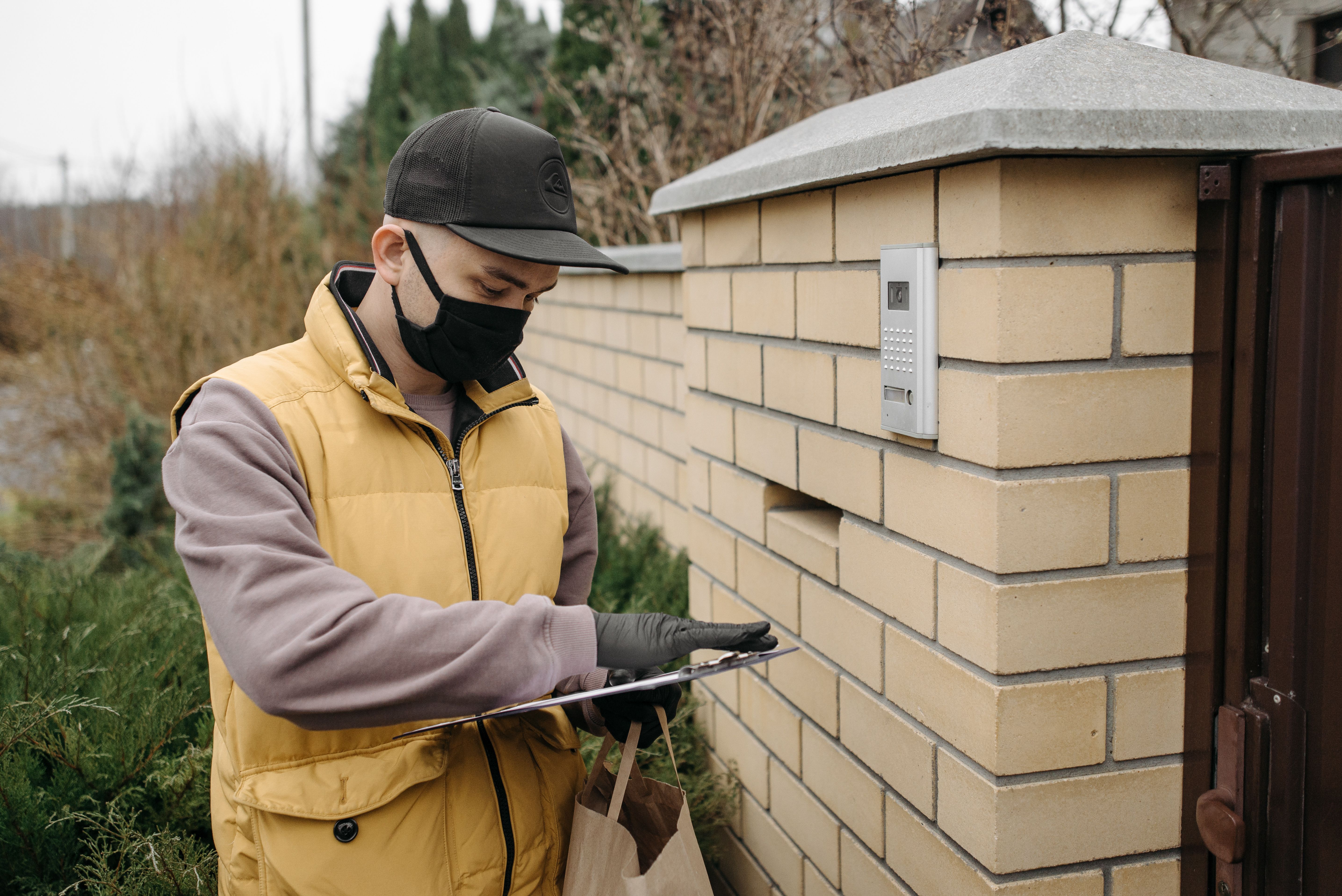 Homem realizando uma entrega usando um dispositivo eletrônico na entrada de uma residência em ambiente externo durante o dia.
