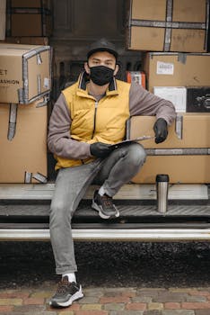 Courier wearing a face mask and gloves sitting with packages, holding a clipboard.