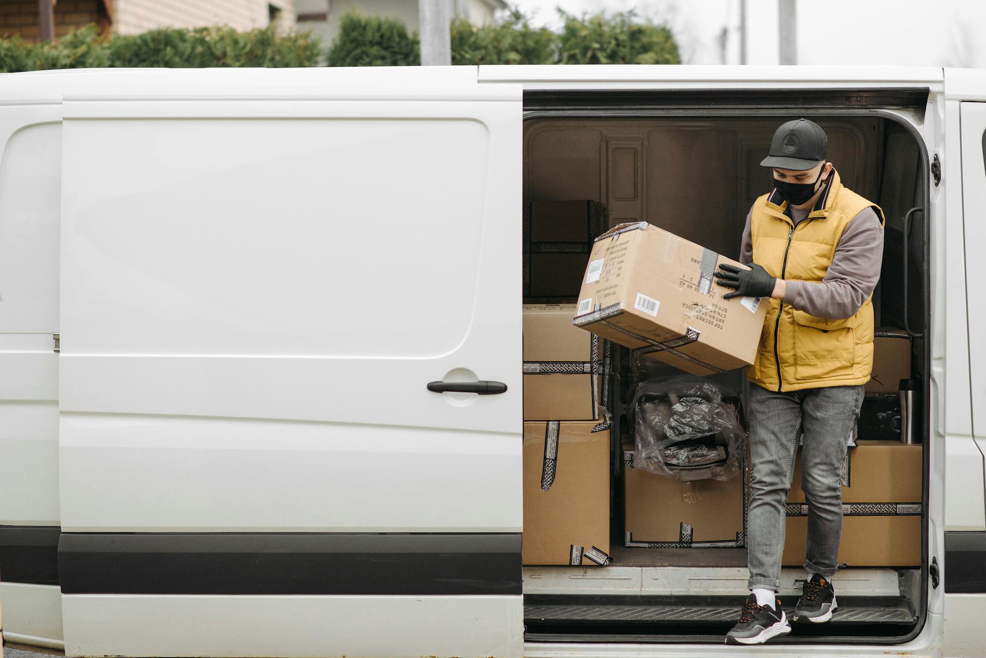 Courier wearing mask unloads packages from white delivery van during the day.