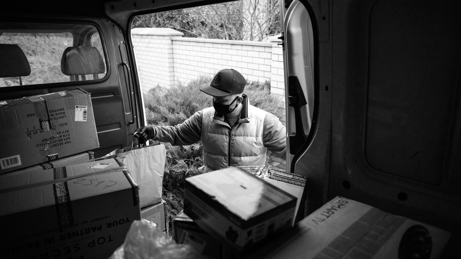 Black and white photo of a masked courier delivering packages from a van loaded with boxes.