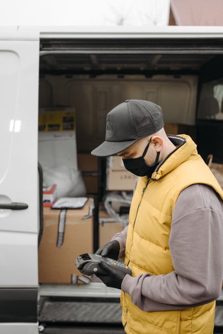 A masked courier handling a payment terminal next to a van loaded with packages, symbolizing safe delivery during the pandemic. A masked courier handling a payment terminal next to a van loaded with packages, symbolizing safe delivery during the pandemic.