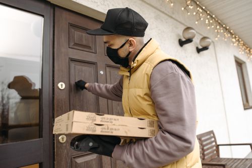 Free A deliveryman wearing a mask and gloves knocks on a door while holding pizza boxes, emphasizing safety during delivery. Stock Photo