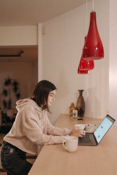 A woman in a hoodie working on a laptop in a cozy home setting with a coffee mug.