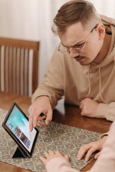 Young man using a tablet with touchscreen indoors, wearing eyeglasses and a hoodie.