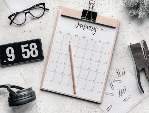 Top view composition of clipboard with calendar and pencil placed on desk amidst stationery and eyeglasses