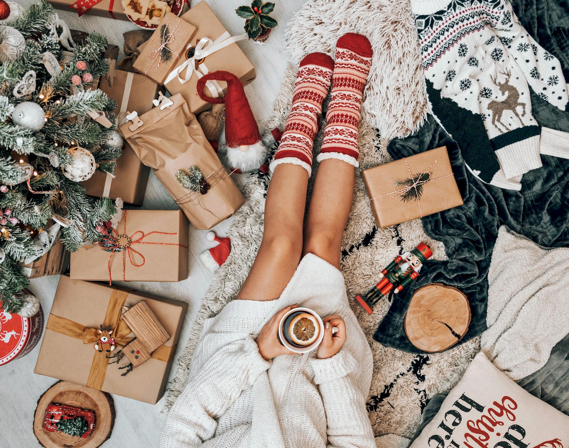 Overhead view of a person sat wearing a wooly cardigan and big Christmassy socks with a cup of mulled cider in their hands. They are surrounded by Christmas presents, tree, and decorations. 