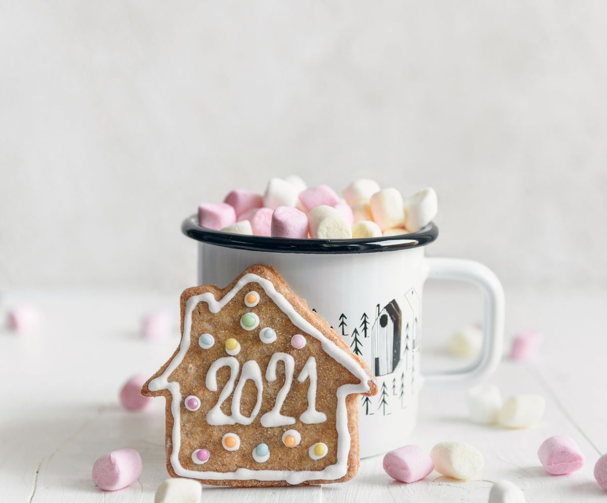 Festive gingerbread cookie with inscription 2021 placed against mug of delicious hot drink garnished with sweet marshmallows and served on white table