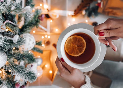 Top view crop anonymous female with manicured hands holding cup of hot fresh tea with lemon while standing near sparkling decorated Christmas tree