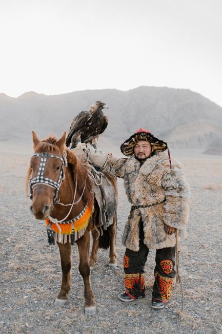 Asian man in fur clothing with horse and eagle in a Mongolian landscape, showcasing traditional culture.