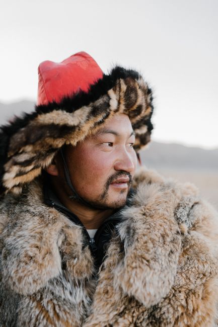 A Mongolian man wearing traditional fur clothes and hat in a serene outdoor setting.