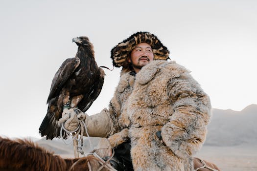 A Mongolian eagle hunter in fur coat with a golden eagle perched on arm in the mountains.