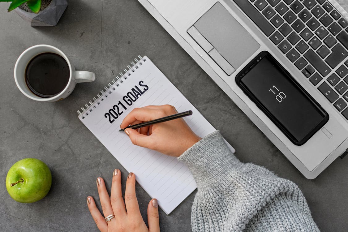 Top view crop anonymous female in sweater writing down goals for 2021 year in notebook while sitting at desk with laptop and cup of coffee