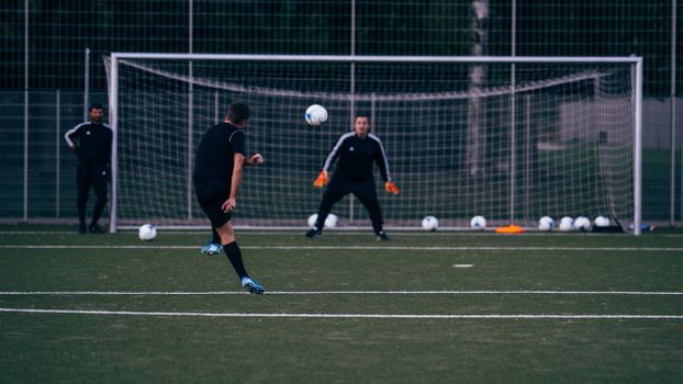 Male football player kicking ball towards goalkeeper standing against goal during training in field