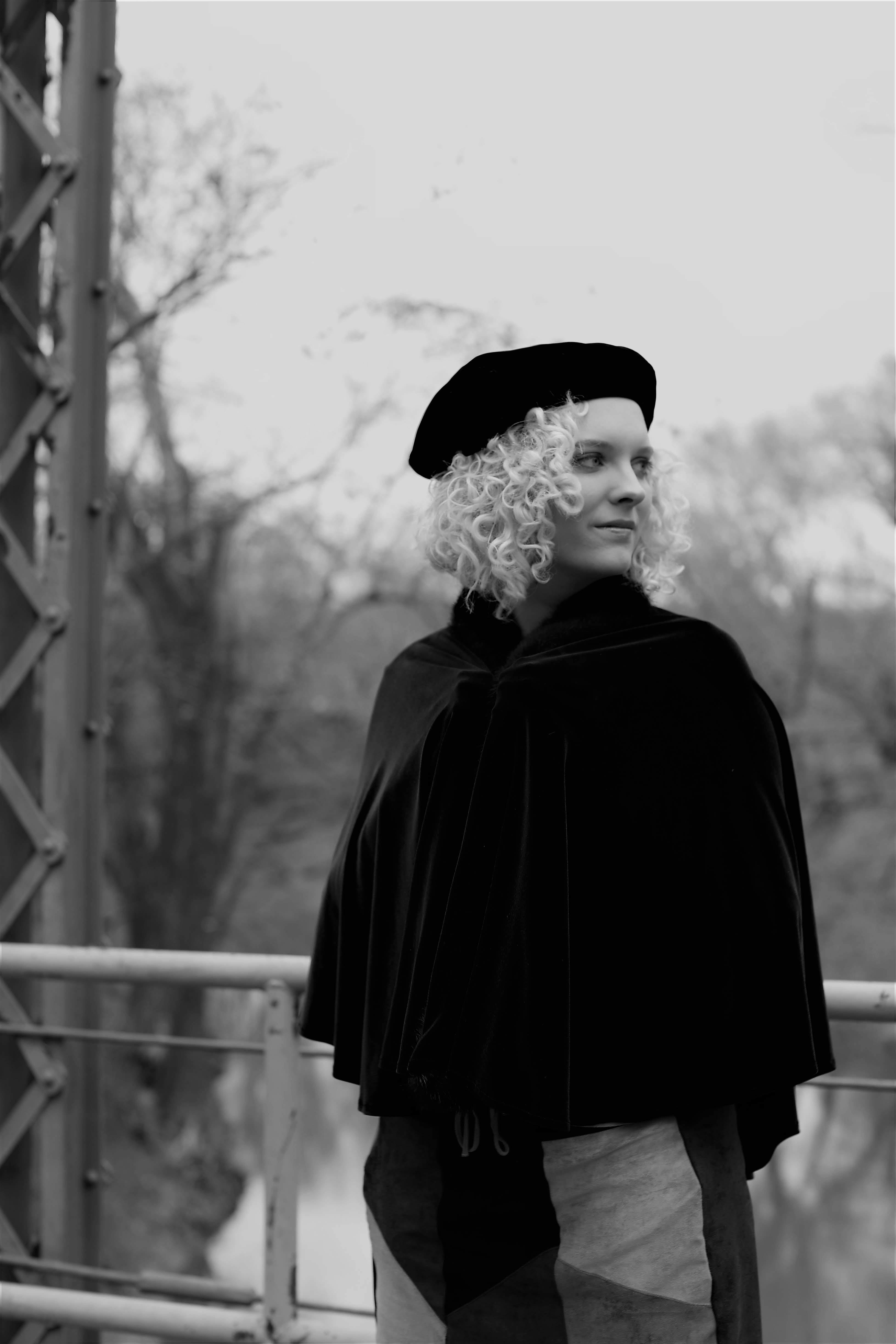 Black and white photo of a fashionable woman in a cape and beret on a bridge, exuding retro style.