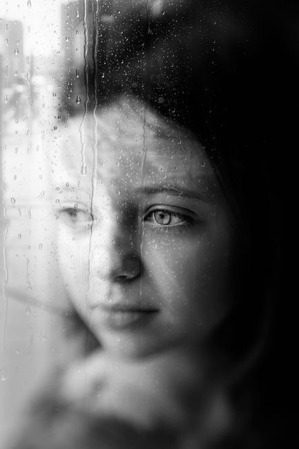 Black and white of little wistful girl staring in window with many raindrops