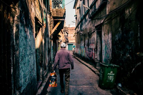 Back view of faceless male in casual clothes carrying plastic package and walking through narrow street