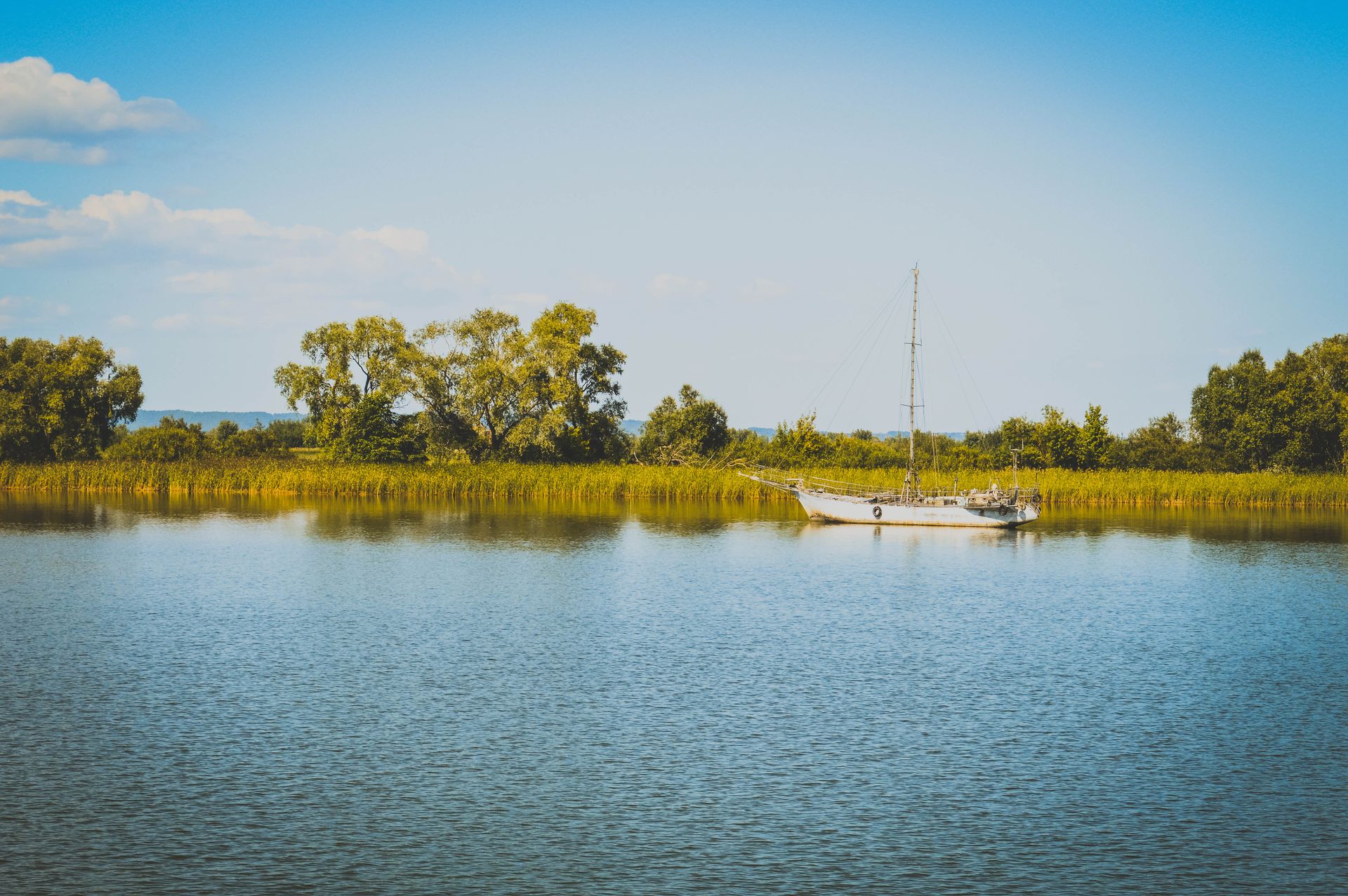 A tranquil scene featuring a lone sailboat on a calm river surrounded by lush greenery and clear blue skies.