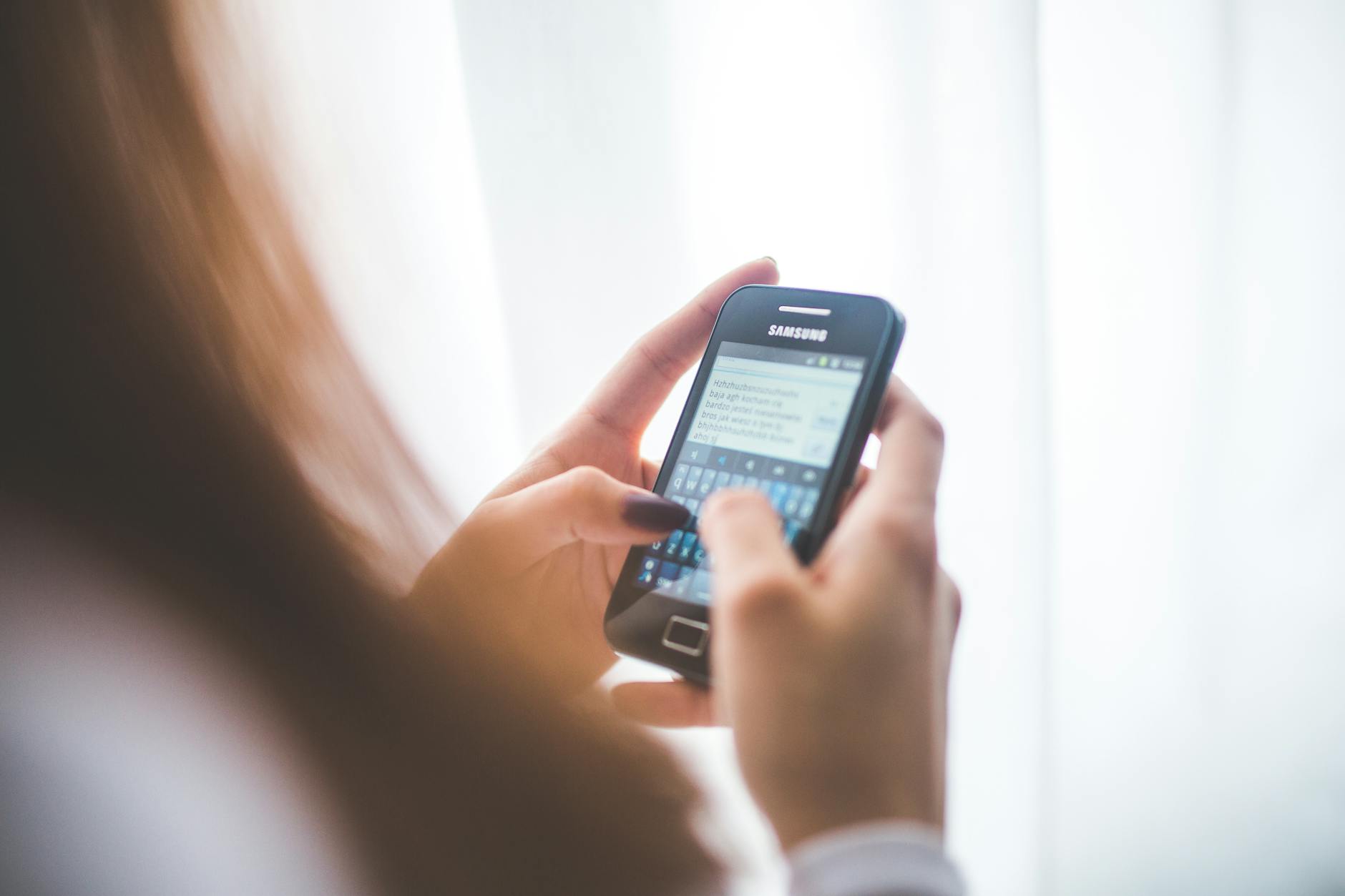 Close-up of a young woman typing on a Samsung smartphone at home. Bright indoor lighting.