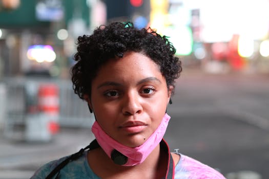 Portrait of a young woman with a pink face mask around her neck in an urban setting with blurry lights.