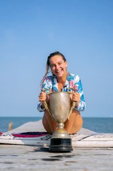A cheerful woman sitting on a boardwalk holding a large trophy, against a clear blue sky.