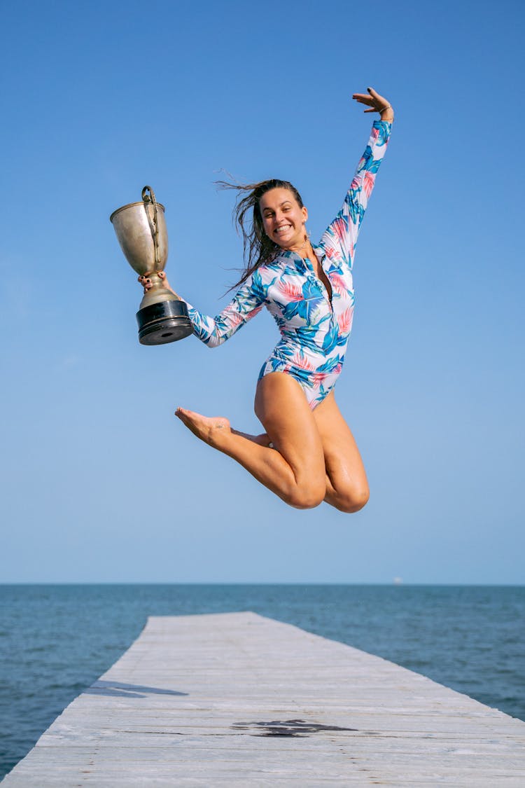 A Happy Woman In Floral Rash Guard Holding A Trophy While Jumping