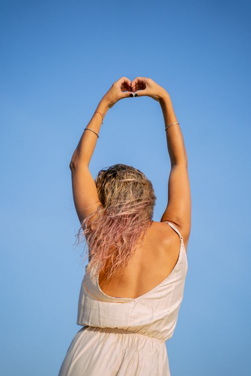 Free Woman in a backless white dress making a heart shape with arms raised against a clear blue sky. Stock Photo