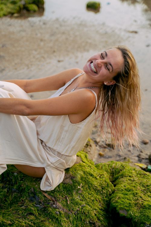 Free Smiling woman in a white dress enjoying a moment on mossy rocks by the sea. Stock Photo