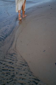 A woman in a skirt and anklet walks barefoot on the sandy beach, leaving footprints in the wet sand.