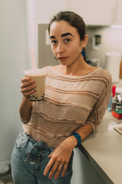 A Woman in Brown Blouse Leaning on the Kitchen Counter while Holding a Plastic Cup of Milk Tea