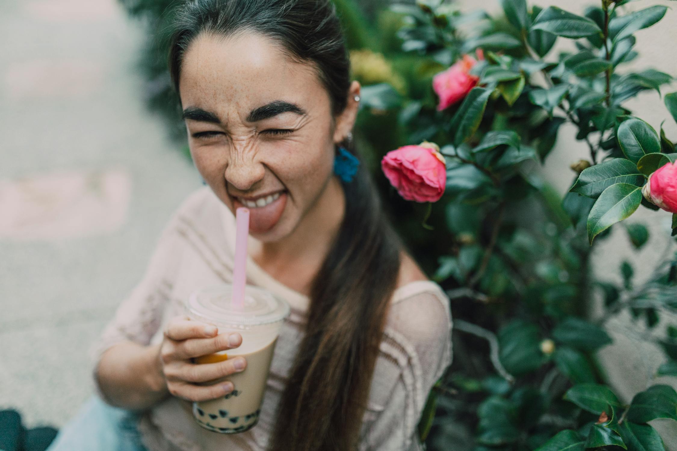 A Woman Holding Cup Of Milk Tea Near Green Plants With Pink Flowers a-woman-holding-cup-of-milk-tea-near-green-plants-with-pink-flowers
