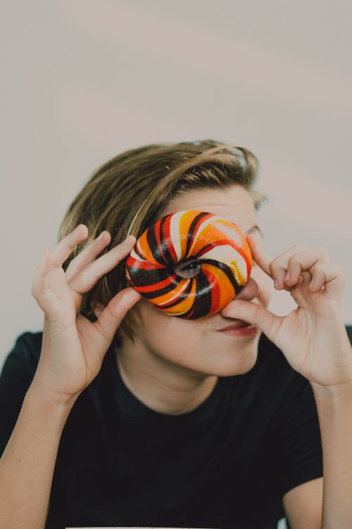 Free A teenager playfully holds a vibrant striped donut up to their eye indoors. Stock Photo