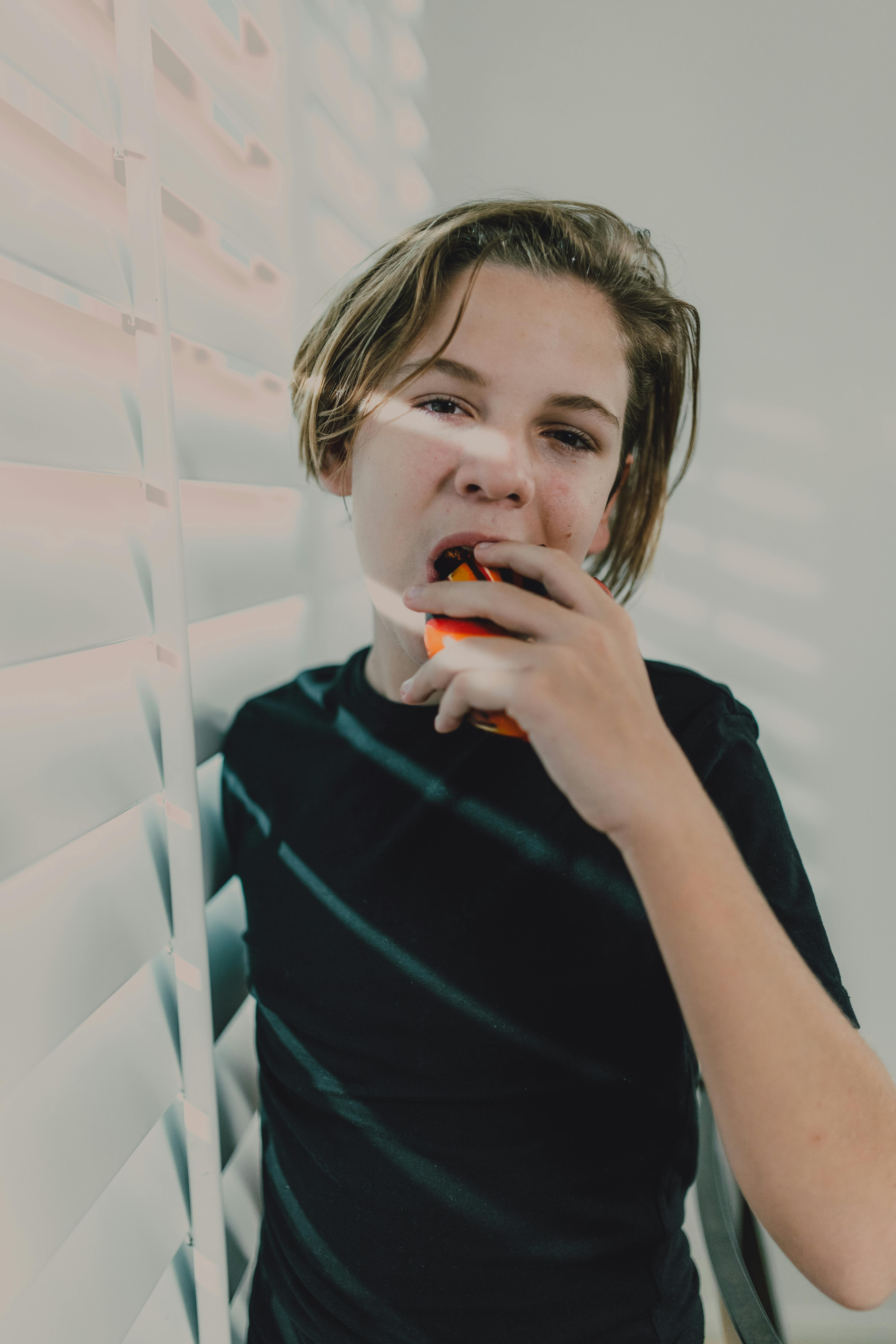 Teenage boy in black shirt enjoying a piece of fruit by window blinds, sunlight creating patterns on him.