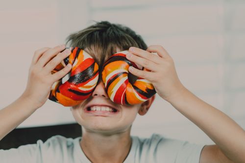 Free Young boy holding vibrant bagels over eyes in playful gesture indoors. Stock Photo