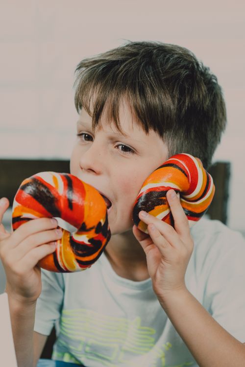 Free Young boy eating vibrant rainbow bagels inside, capturing a playful snack moment. Stock Photo