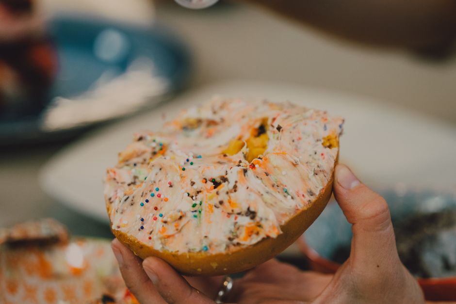 A delicious bagel topped with colorful cream cheese and sprinkles held in hand.