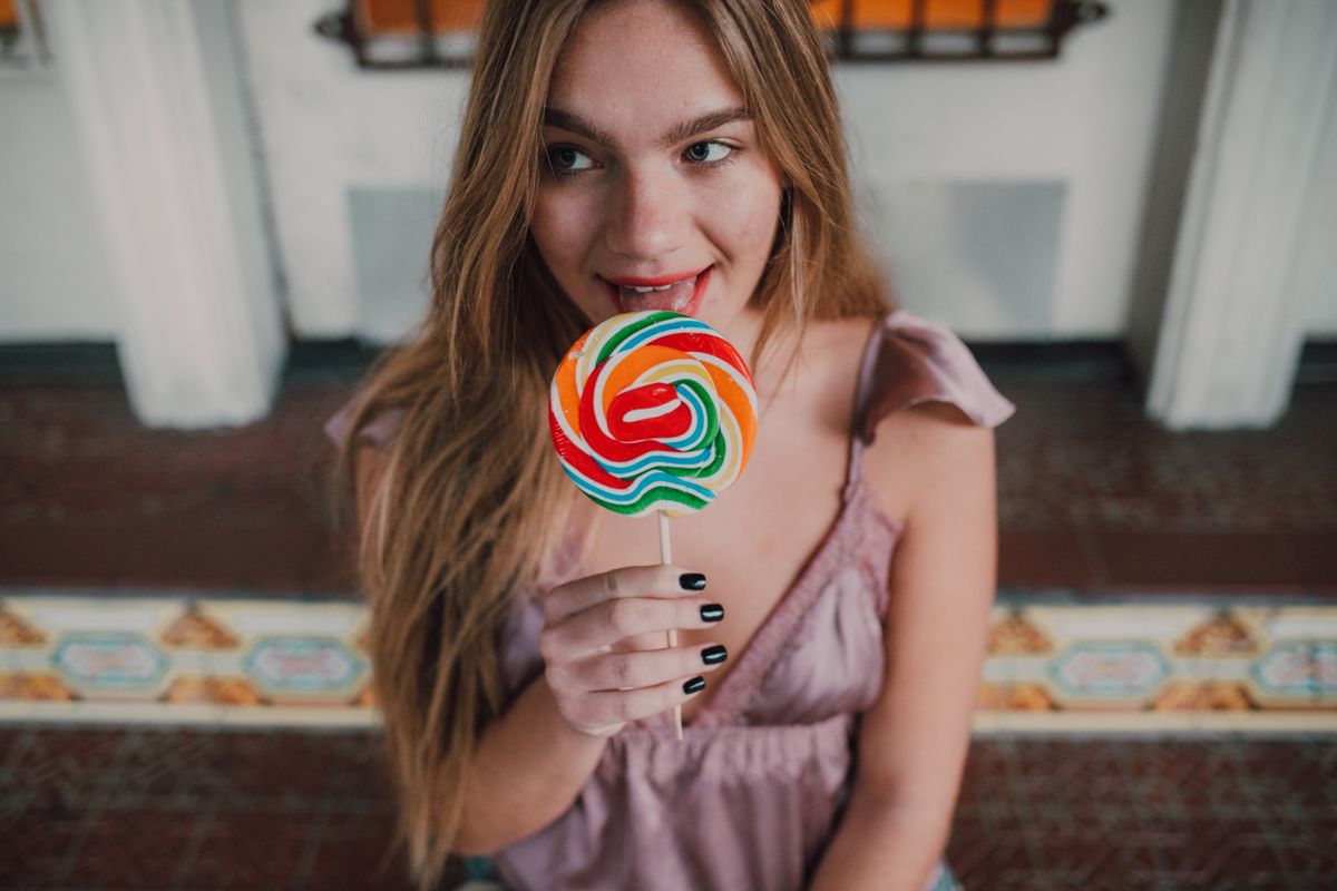 A young woman with long hair enjoys a vibrant lollipop indoors, exuding youthful joy and style.