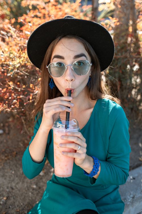 Free Woman wearing sunglasses and hat drinks smoothie outdoors. Perfect summer fashion vibe. Stock Photo