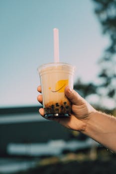 Hand holding a plastic cup of bubble milk tea with straw outdoors, clear sky behind.
