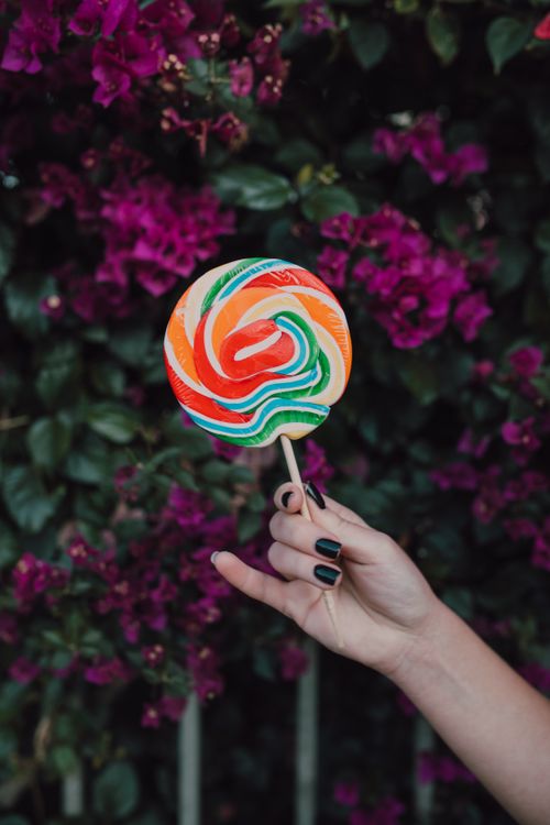 Free Hand holds a rainbow lollipop with vivid flowers in the background, creating a striking contrast. Stock Photo