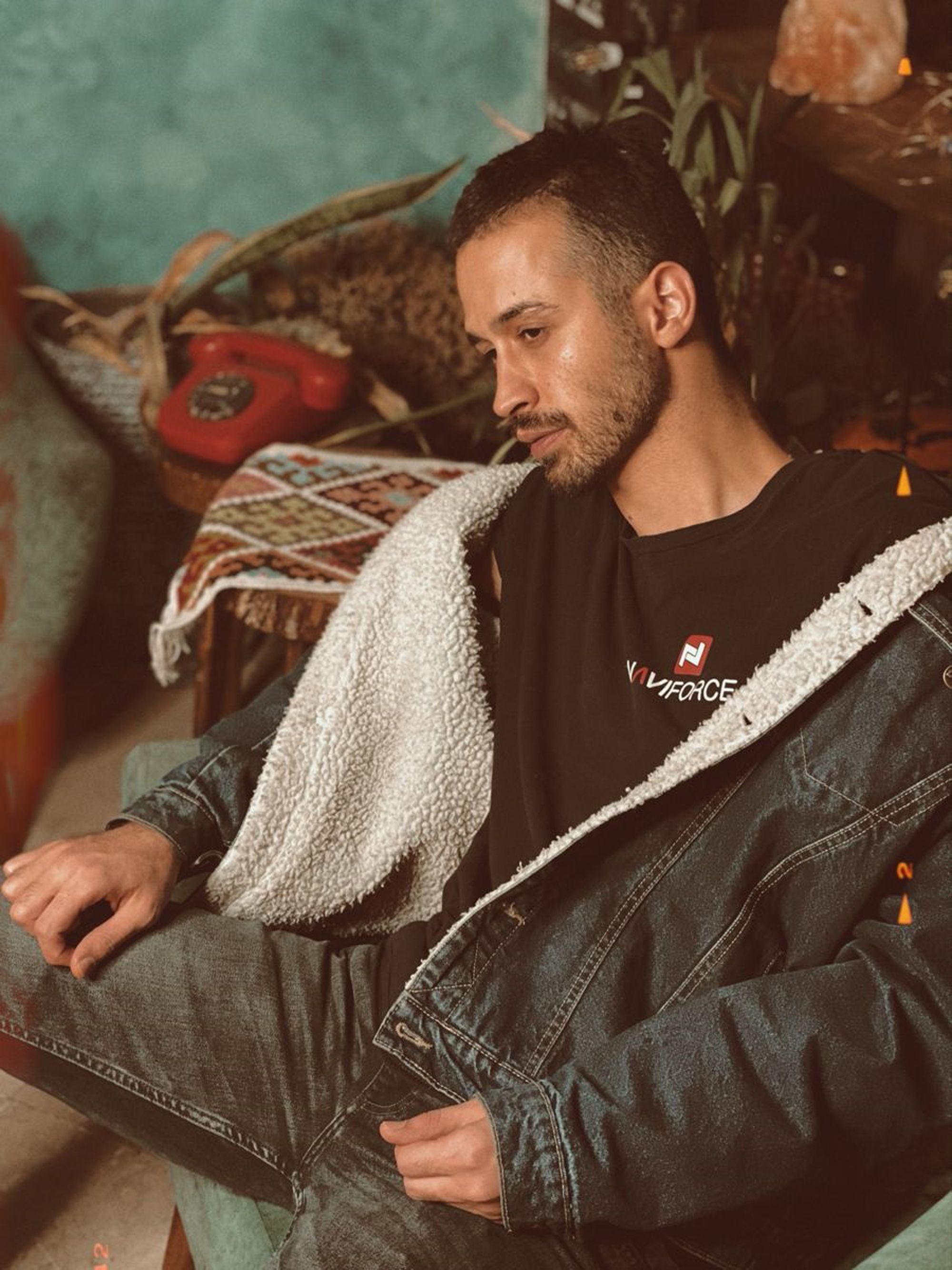 Relaxed man in denim jacket and jeans sitting indoors, looking thoughtful.