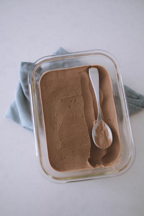 Top view of yummy homemade chocolate ice cream in glass container placed on white table with silver spoon in kitchen