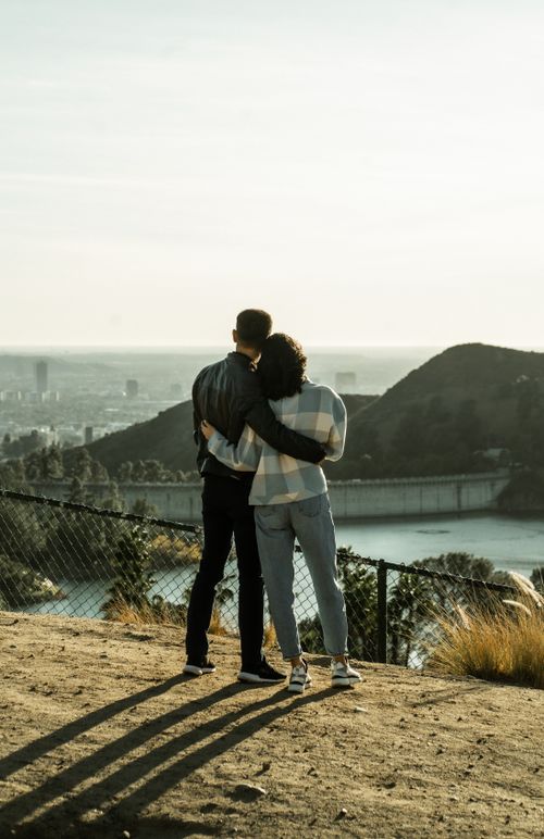 Free Back view of full length unrecognizable couple in casual clothes standing on bank of reservoir hugging and admiring view Stock Photo