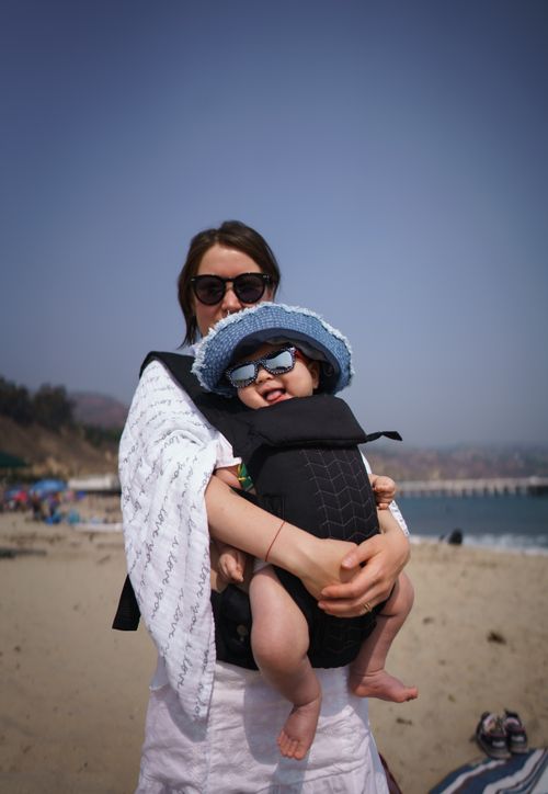 Free Young mother with adorable smiling baby in sunglasses and hat in baby carrier on sandy beach in sunny weather looking at camera Stock Photo