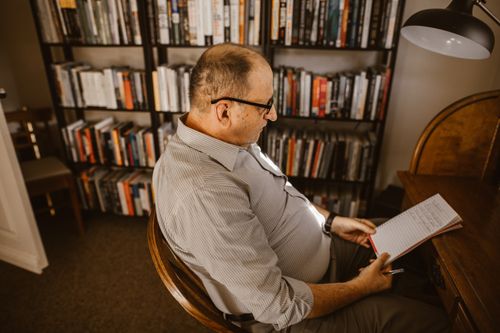 Free An elderly man reading and writing in a cozy home library, surrounded by bookshelves. Stock Photo
