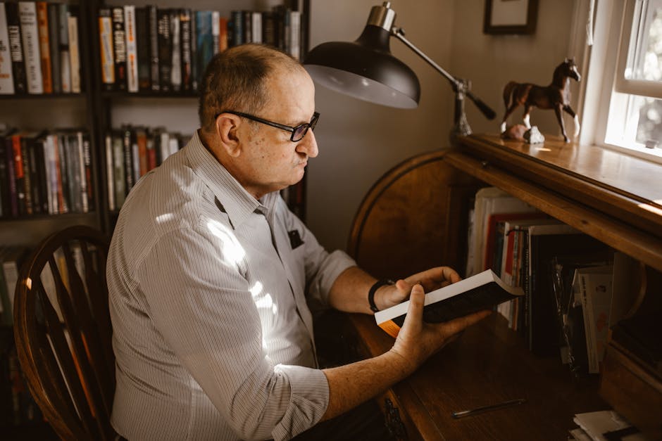 Elderly man in glasses reading a book at a wooden desk with bookshelves and lamp.